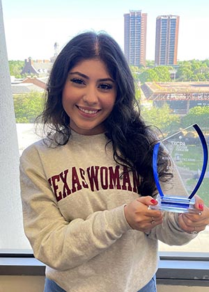 Dayssy Moreno, wearing a TWU sweatshirt, holds a glass plaque in front of a window overlooking the TWU campus