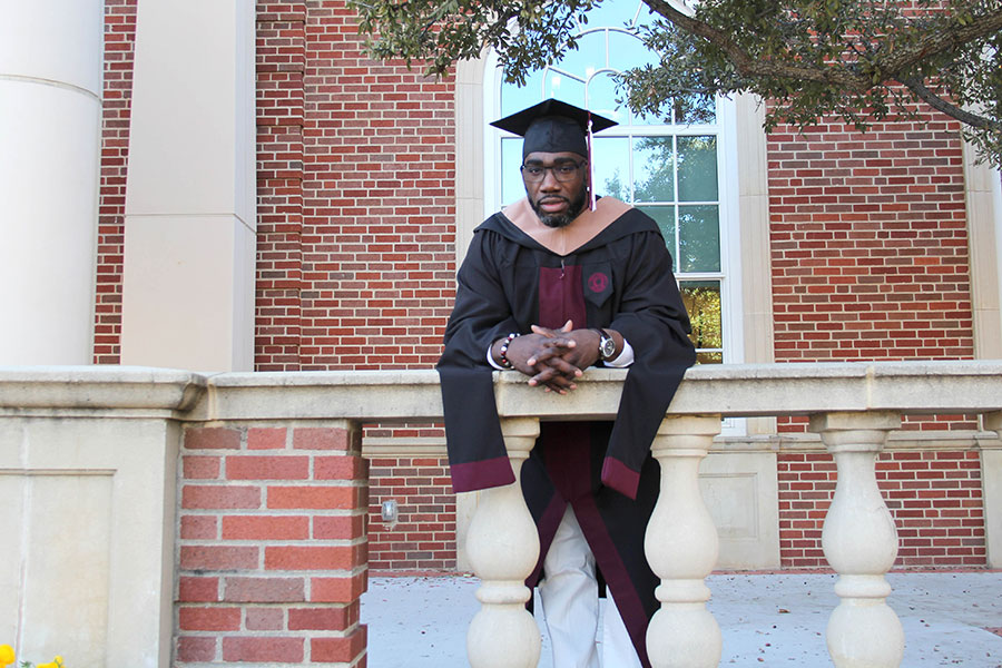 graduate in cap and gown leans on railing in front of brick building