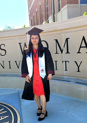 graduate in cap and gown stands in front of Texas Woman's University sign