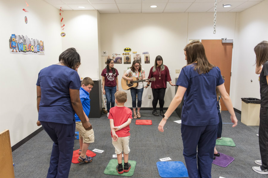 A woman playing the guitar to a group of dancing kids in the clinic with nurses around.