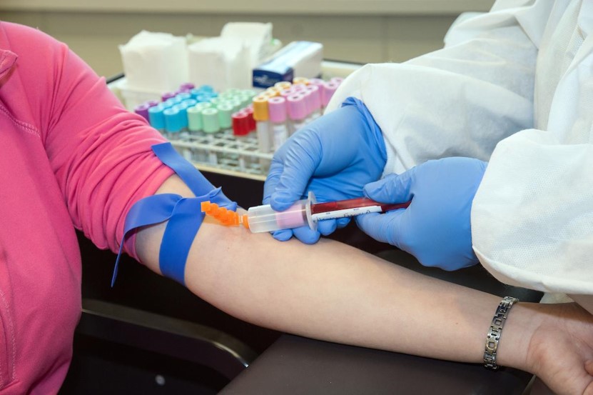 A person with gloved hands collecting blood with a tube from a person with their arm extended.