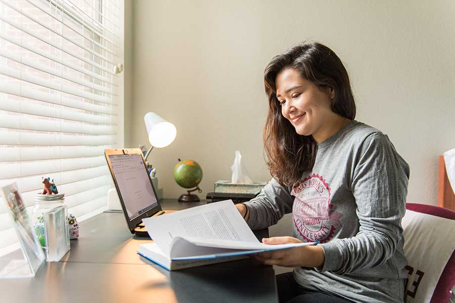 A TWU student works at a desk in her bedroom with a laptop and book in front of her.