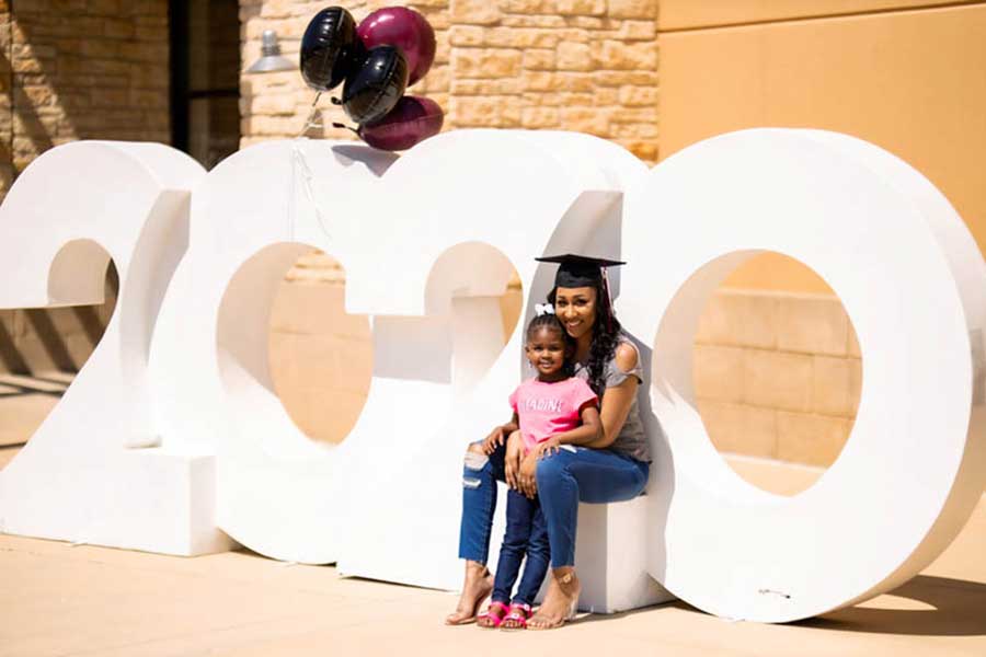 A TWU graduate sits on a large 2020 lettering with her daughter on her lap.