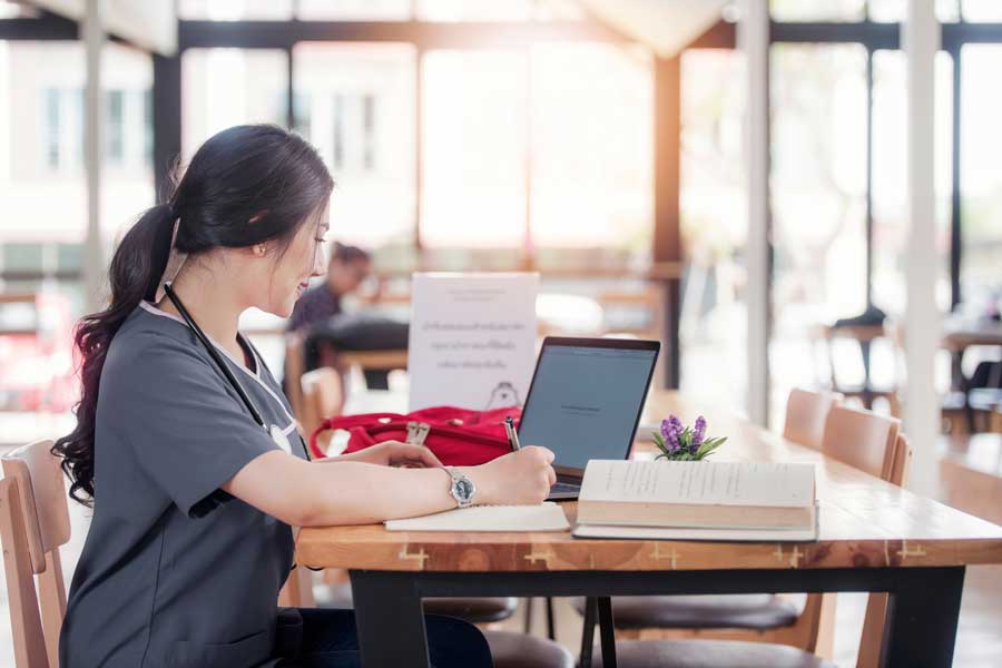 A health sciences student in medical scrubs works on a laptop.