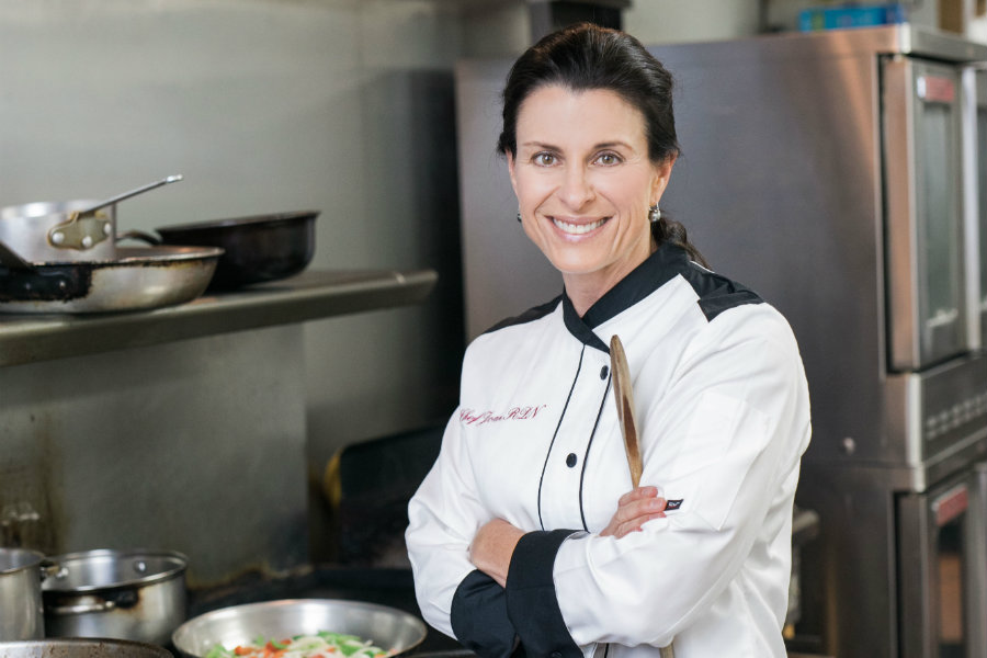 Joan Denton in a professional chef uniform and posing in a professional kitchen setting with food cooking on a stove.