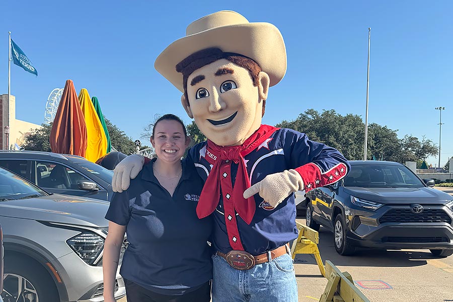 a TWU student in a blue State Fair polo stands next to a person dressed as Big Te