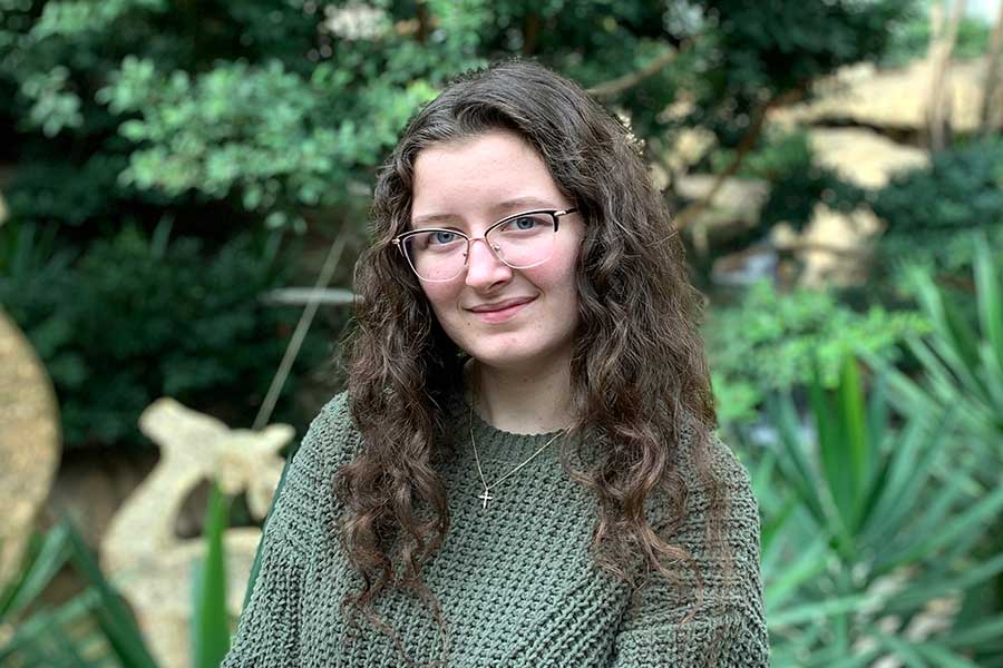 a student in green sweater stands in front of trees and bushes