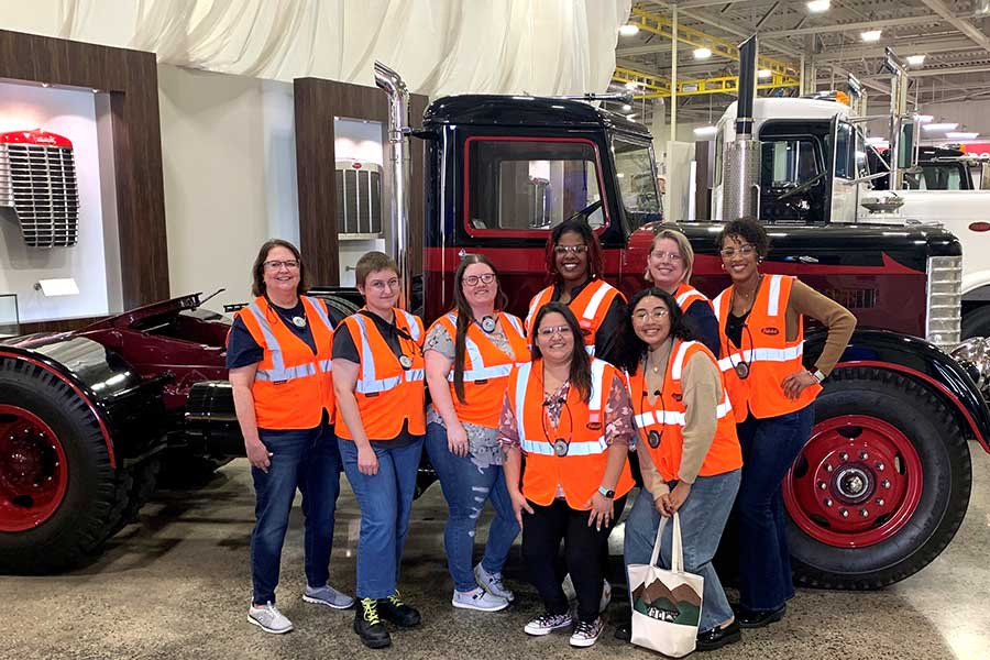 seven students and faculty member in bright orange vest stand in front of a large truck inside a facility