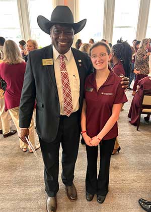 student in maroon polo stands next to man in suit in a ballroom 