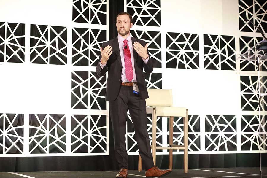 man in black suit with red tie stands on stage in front of black and white backdrop