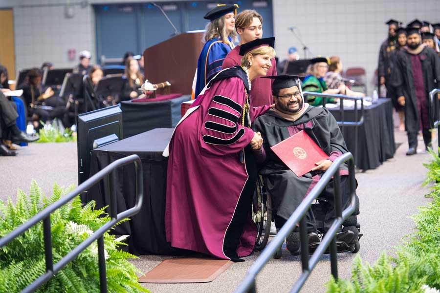 A graduate in academic regalia goes across the stage in a wheelchair during commencement.