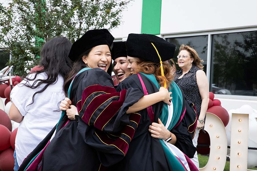 Three graduate students hug enthusiastically in their academic regalia
