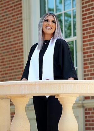 a student in graduation attire stands behind a balcony and in front of a brick building 