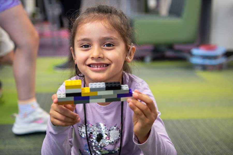 young girl in purple shirt holds up LEGOs while sitting in a carpeted room.