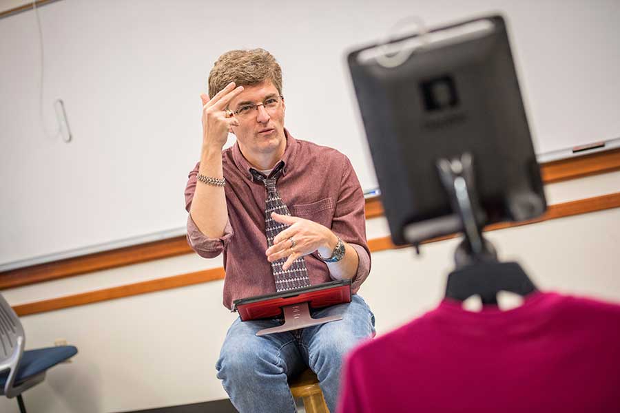 a professor, sitting on a stool, uses his hands to sign to a computer
