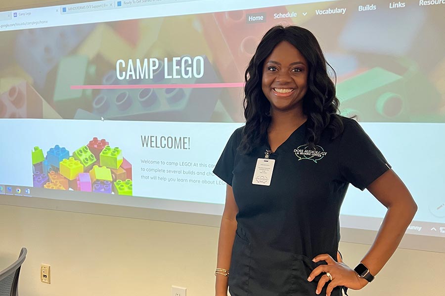 a woman dressed in black scrubs stands in a classroom with a Camp Lego video monitor in the background