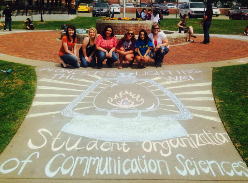 Women sitting in front of chalk art on sidewalk that reads Student Organization of Communication Sciences.	