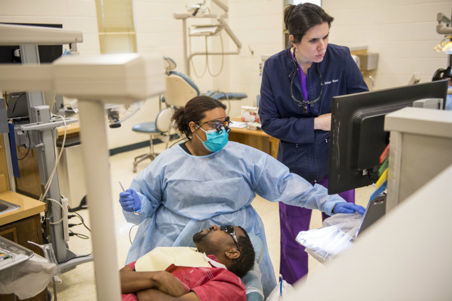 A dental hygiene student working with a patient in a dentist's chair while a professor watches over.