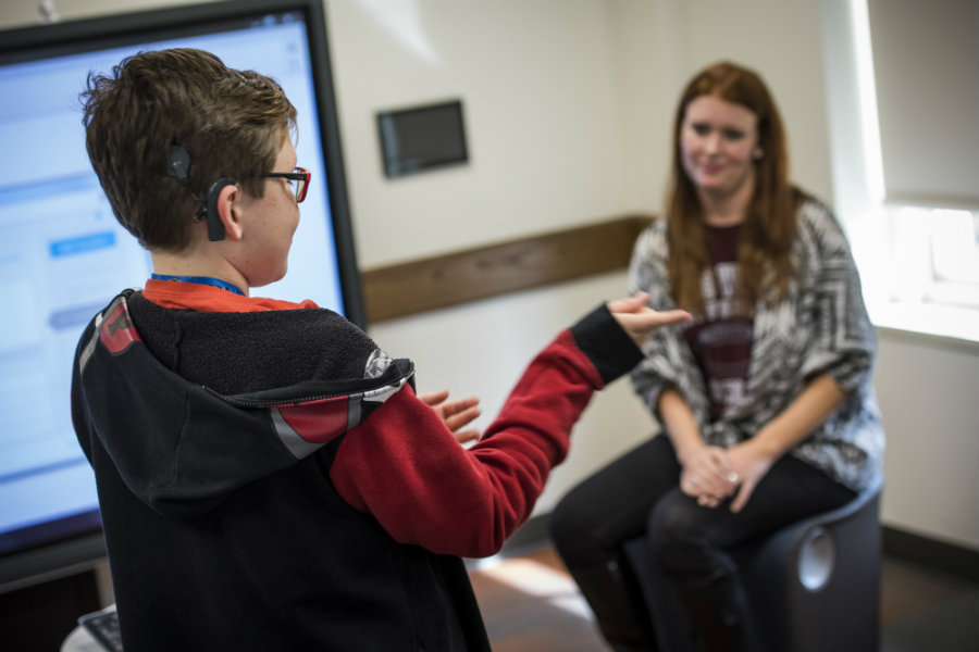 A young boy wearing a hearing aid speaks in sign language to a TWU student in the background.