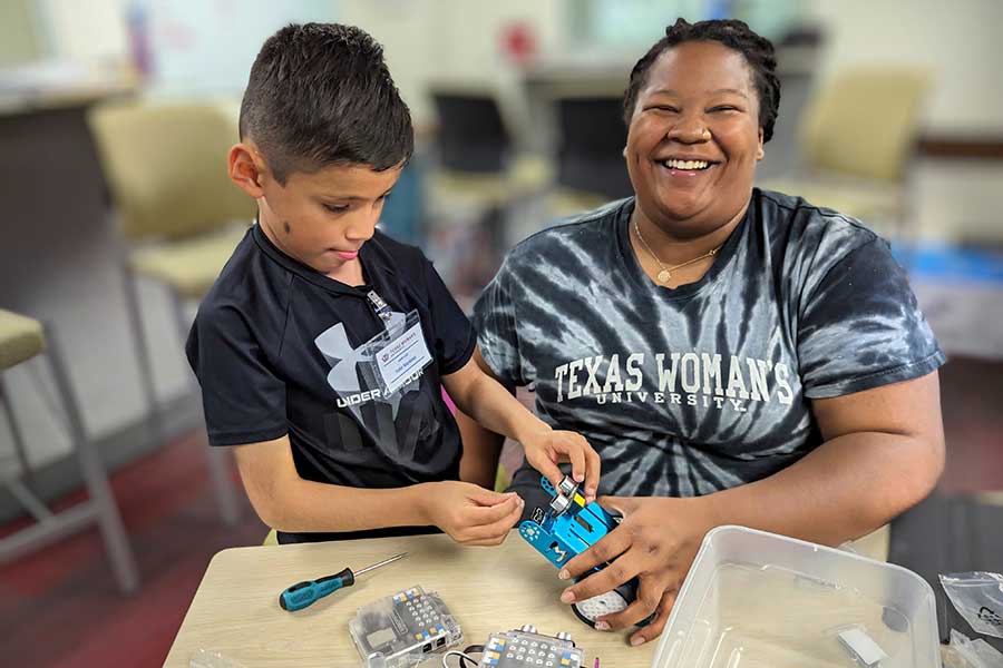 staff member smiles at student working on robot on a table.