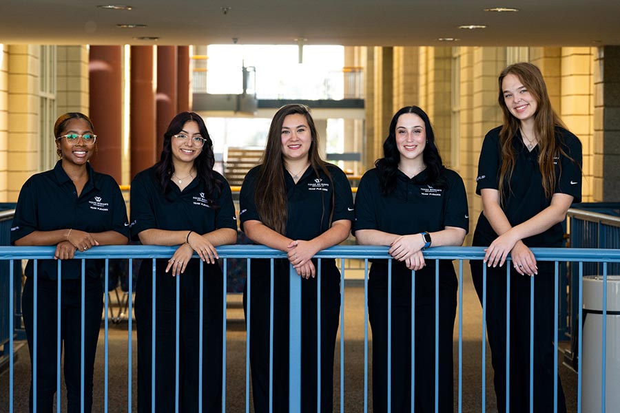 five female kinesiology students dressed in black, stand behind a blue railing.