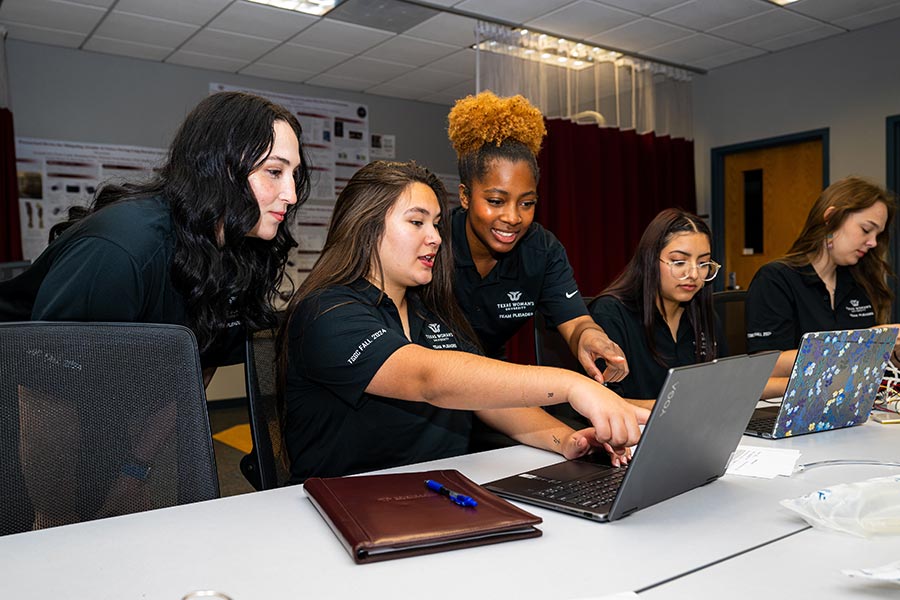 five students in black polos work at a large table with a laptop