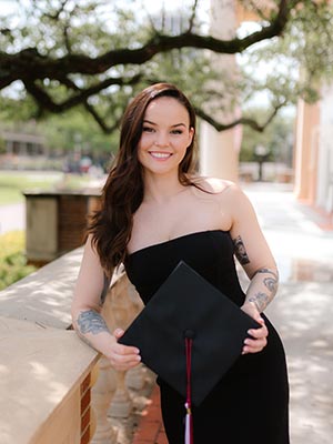 a TWU graduate leans against a TWU building holding a graduation cap