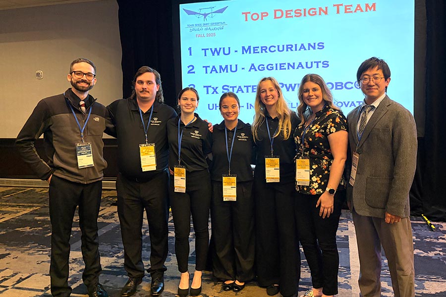 four students in black polos stand with three staff members in front of large display