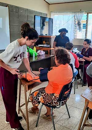 student in TWU gray shirt checks blood pressure of a woman sitting at a desk