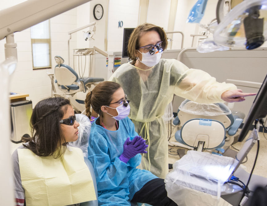 Three women wearing scrubs looking at computer screen with health clinic machinery in background