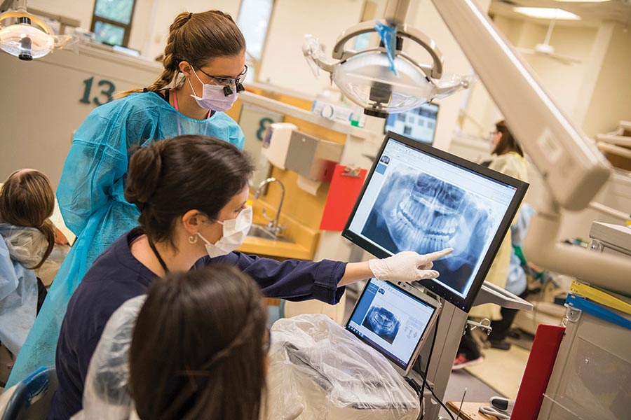 a health professional in blue scrubs points at monitor with x-rays on it while a student in blue scrubs looks on