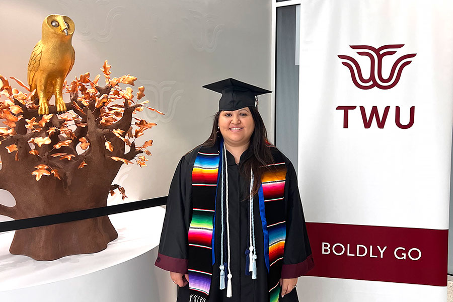 student in graduation regalia stands next to statue of golden owl 