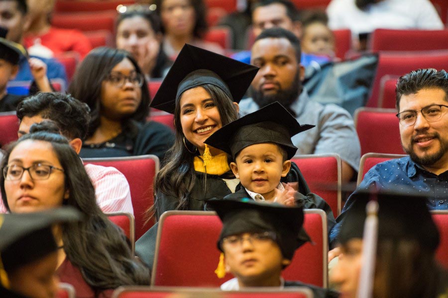 A TWU graduate in academic regalia with her son on her lap in a cap and gown.