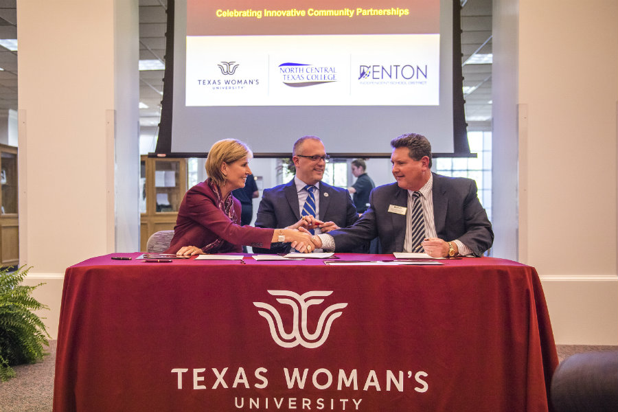 Female in TWU jacket shakes hands with dark-haired man while another man in dark suit sits between t