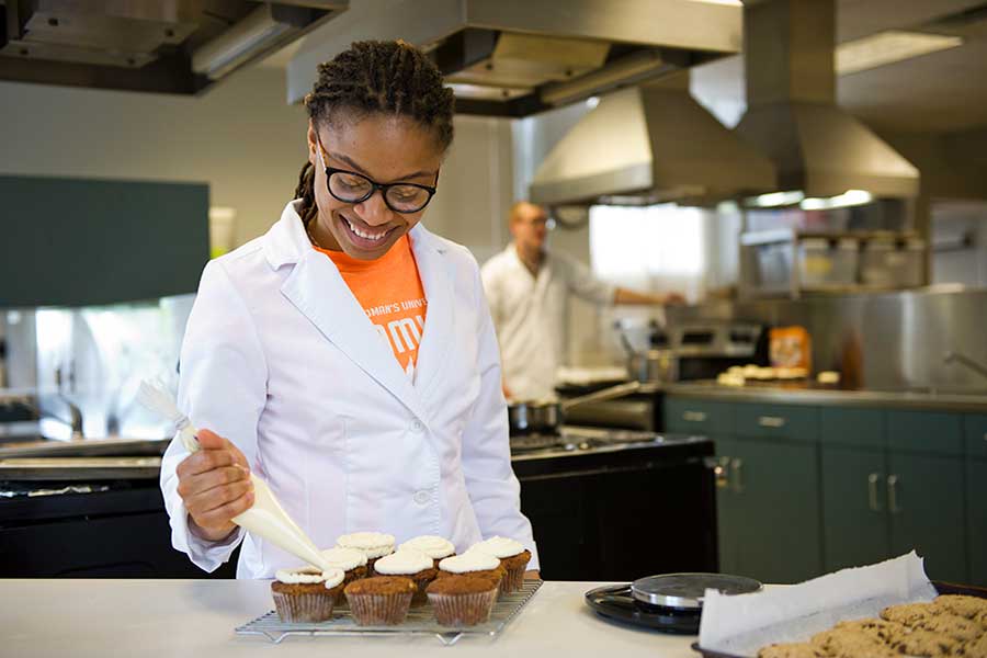 A TWU student ices cupcakes in an industrial kitchen setting.