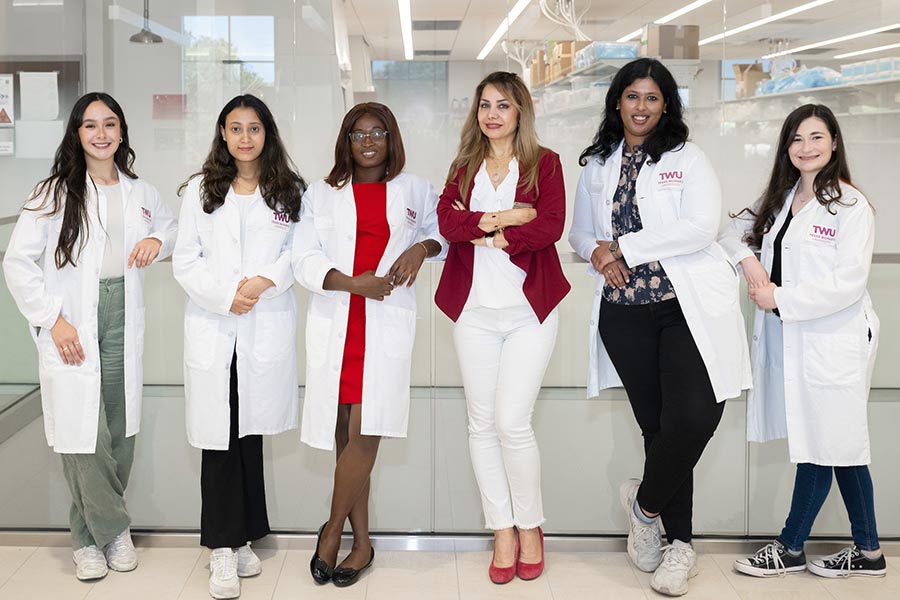 Nutrition professor stands in front of lab with five students next to her in white lab coats