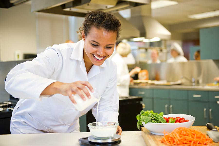 a TWU student in white cooking uniform pours water into bowl in an industrial kitchen setting