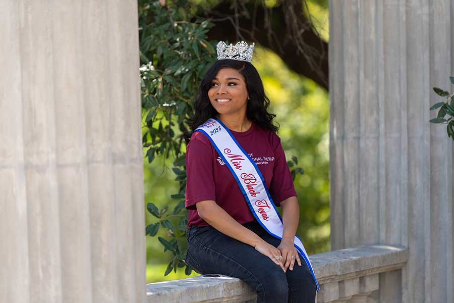 student in maroon shirt sits on ledge with crown and sash