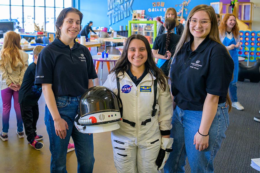 Alyssa Flores stands in an astonaut space suit in between two kinesiology students 