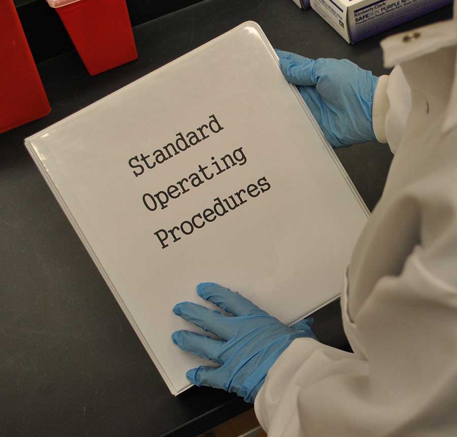 A person with lab coat and blue nitrile gloves holding a white binder with black lettering reading 'Standard Operating Procedures'