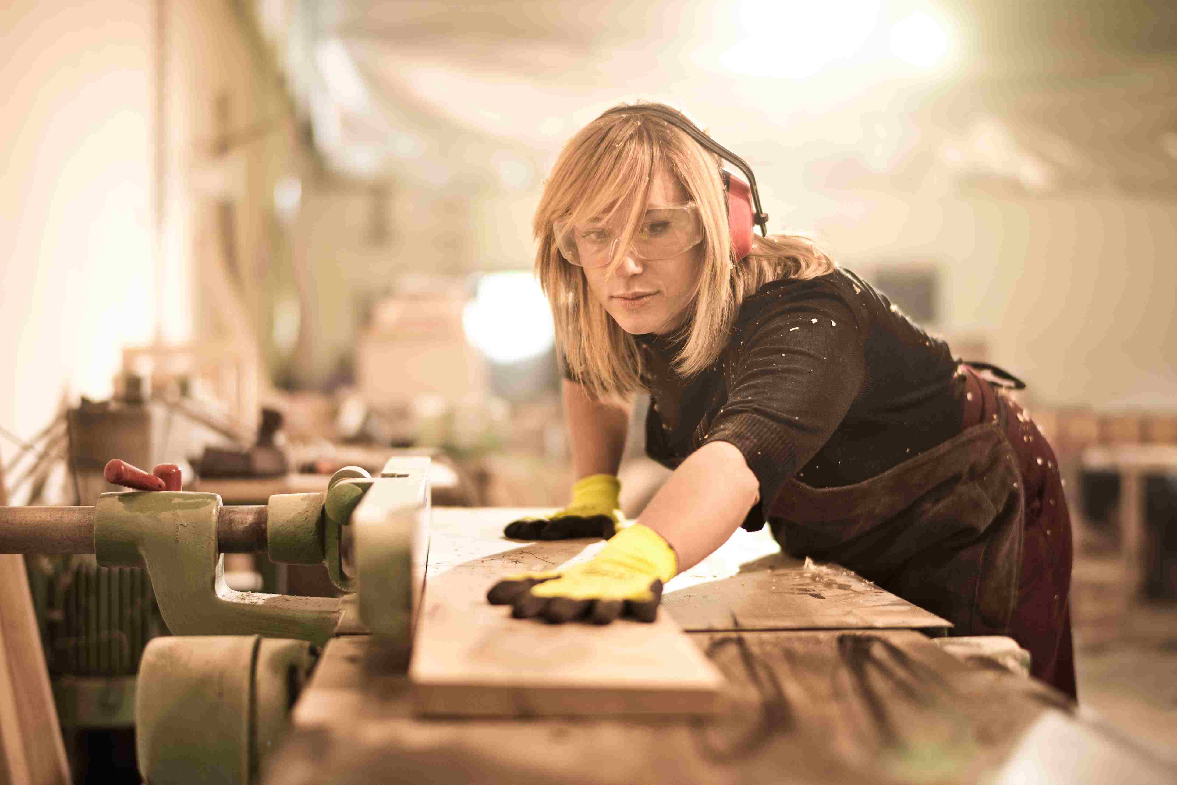 Woman with safety glasses and ear protection guiding a plank of wood through a woodworking machine.