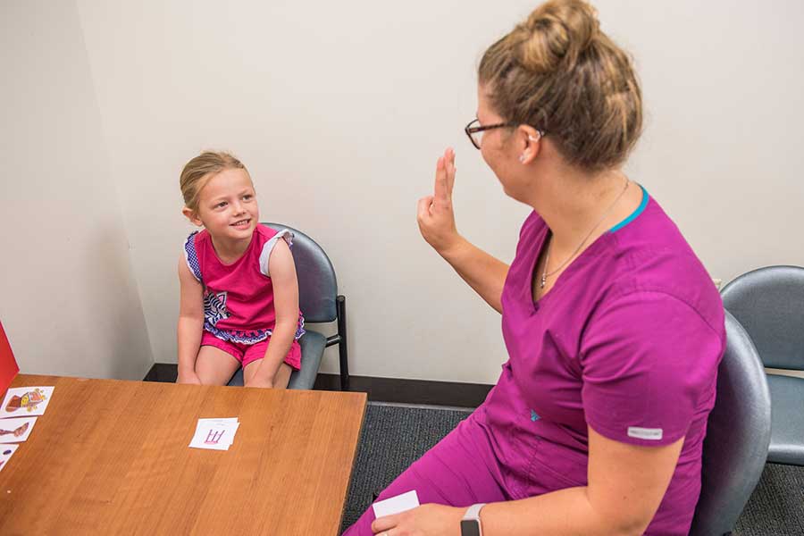 a speech language pathology student in purple scrubs holds up one hand facing a child sitting on a chair