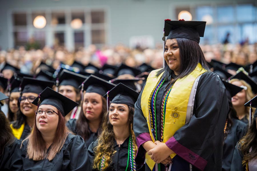 A female student wearing a yellow stole stands apart from the commencement crowd.