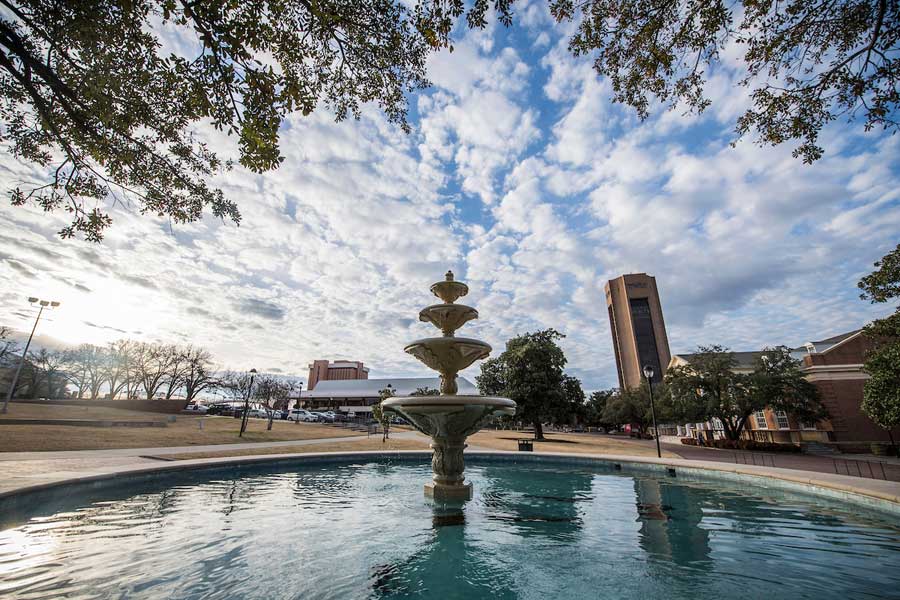The TWU Fountain in the center foreground, with a cloudy sky, ACT and the Student Union in the background	