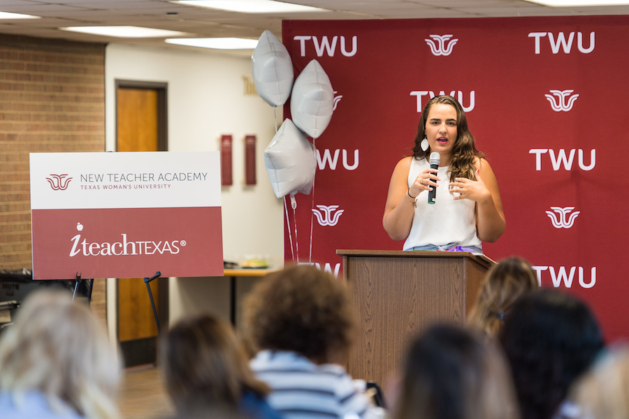Young woman standing in front of TWU banner holds microphone and speaks to audience. A New Teacher Academy/iteachTEXAS sign is to her right.