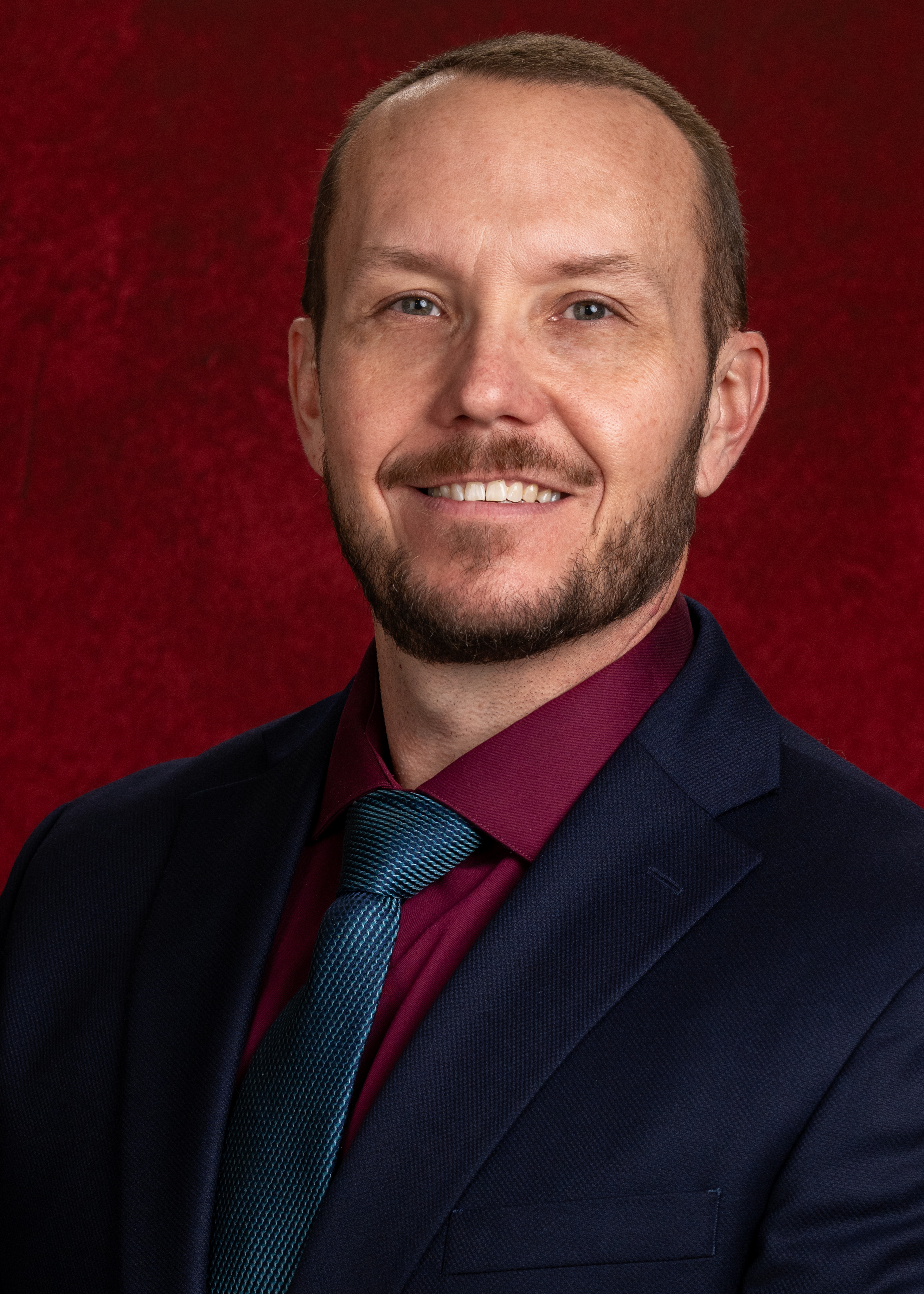 Professional headshot of Allen Claytor wearing business attire, facing the camera with a dark maroon colored background
