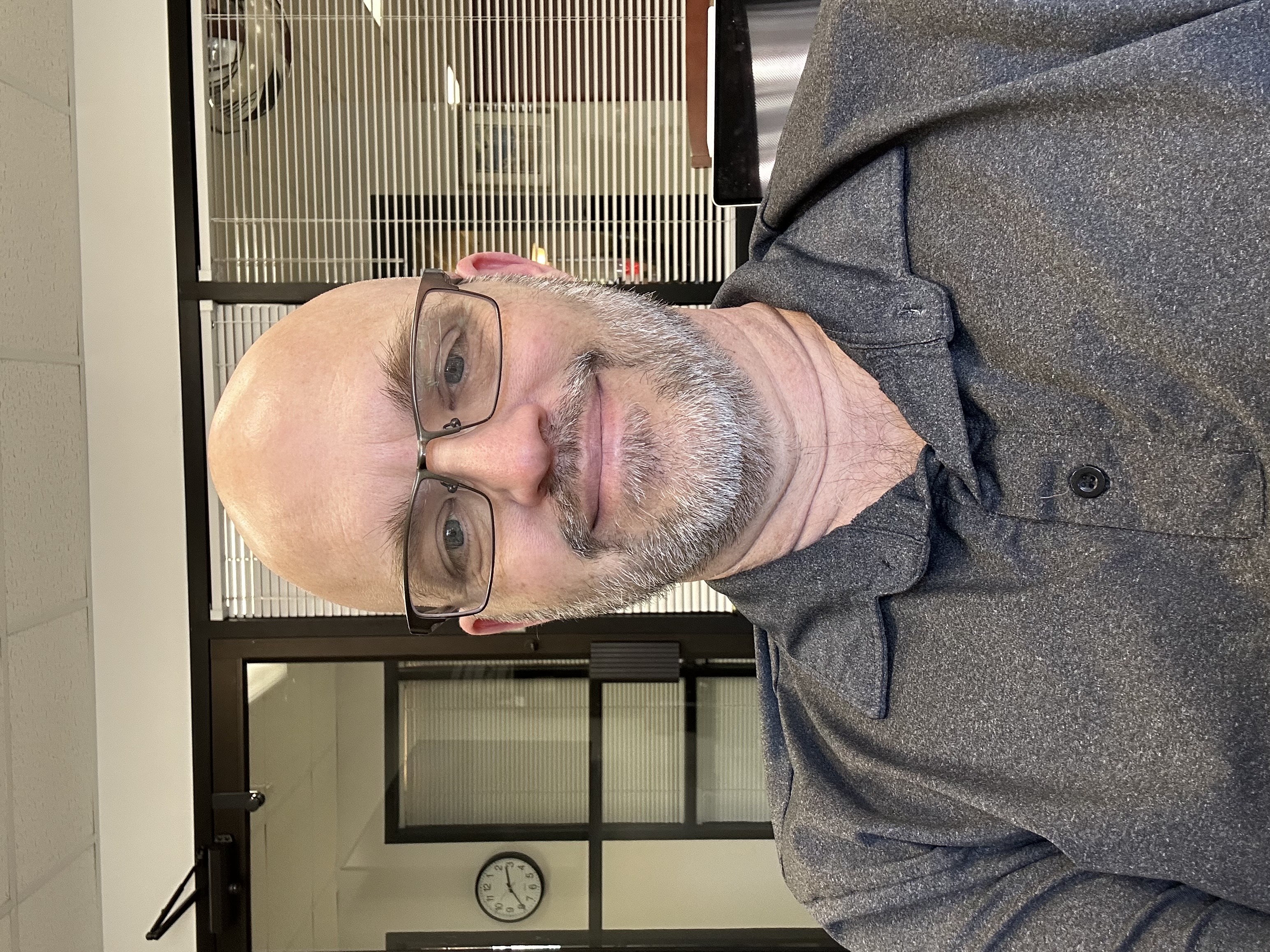 Headshot of AJ Galatas, smiling in an office setting with glass partitions and a clock in the background